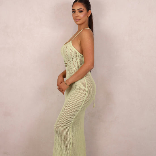 A model wearing a pale green crochet knit maxi dress with a floral waist cut-out and open criss-cross back.