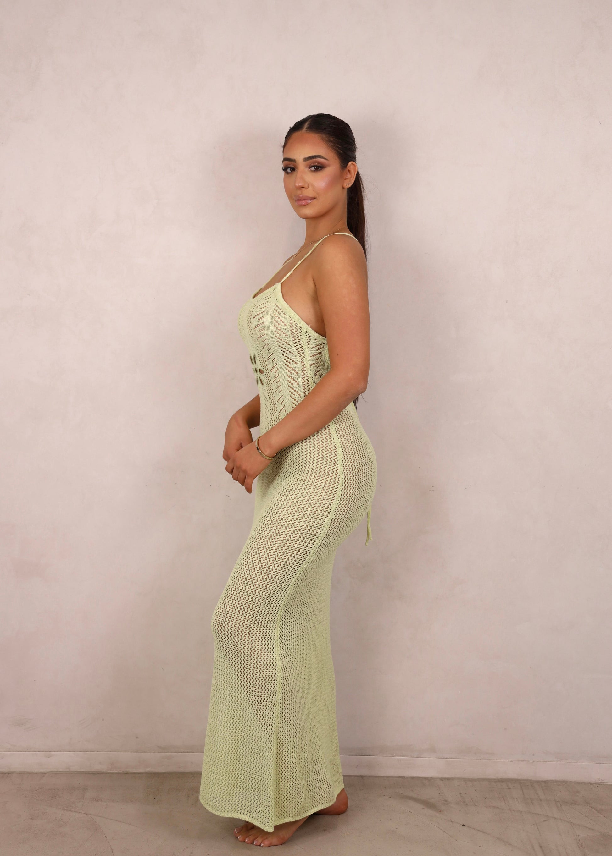A model wearing a pale green crochet knit maxi dress with a floral waist cut-out and open criss-cross back.