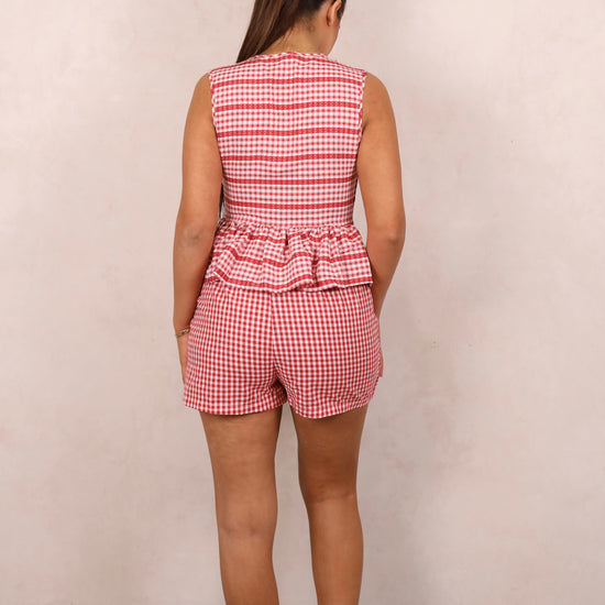 A model wearing a red and white gingham tie-up peplum top and matching wrap front mini skirt, standing against a plain background.