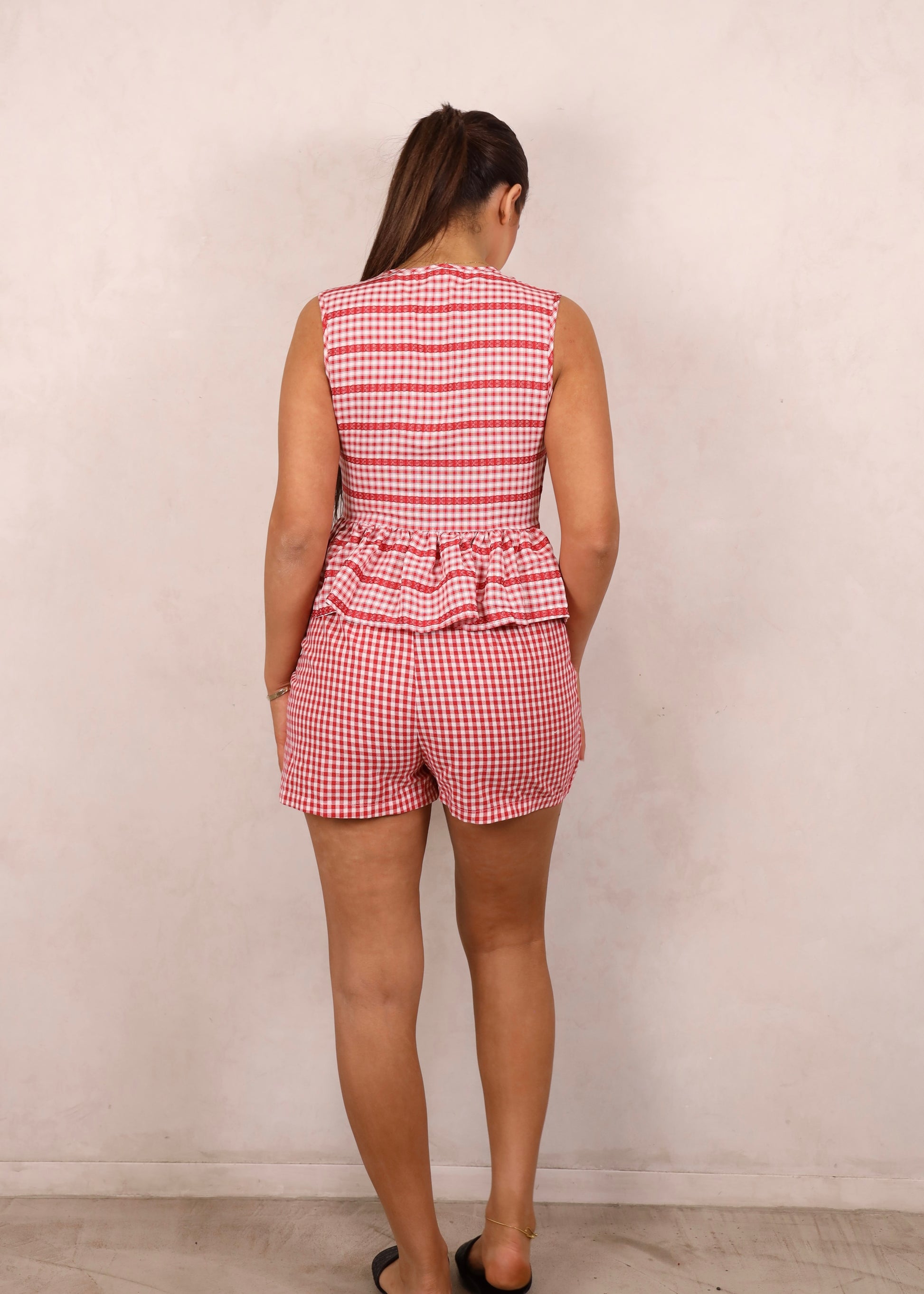 A model wearing a red and white gingham tie-up peplum top and matching wrap front mini skirt, standing against a plain background.
