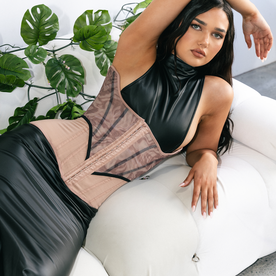 Woman wearing a black and beige corset sitting on a white couch with green plants in the background.
