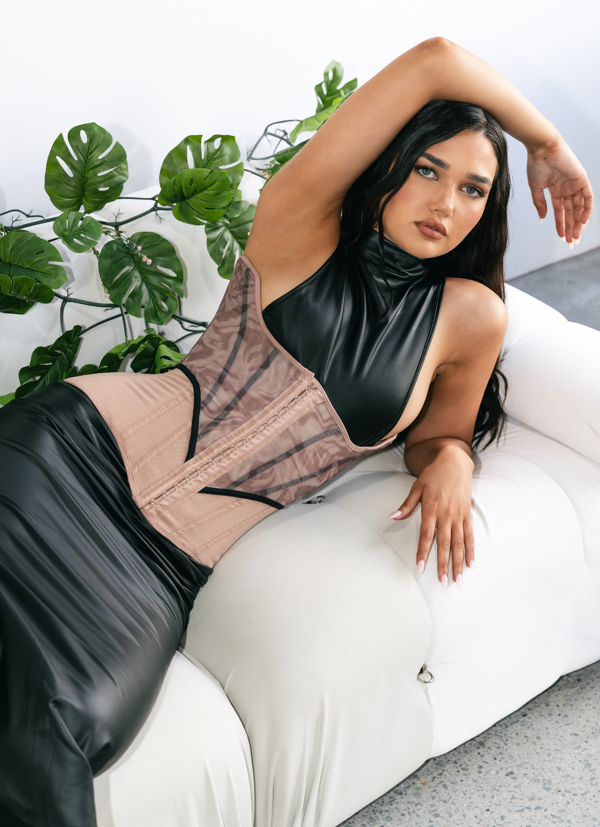 Woman wearing a black and beige corset sitting on a white couch with green plants in the background.